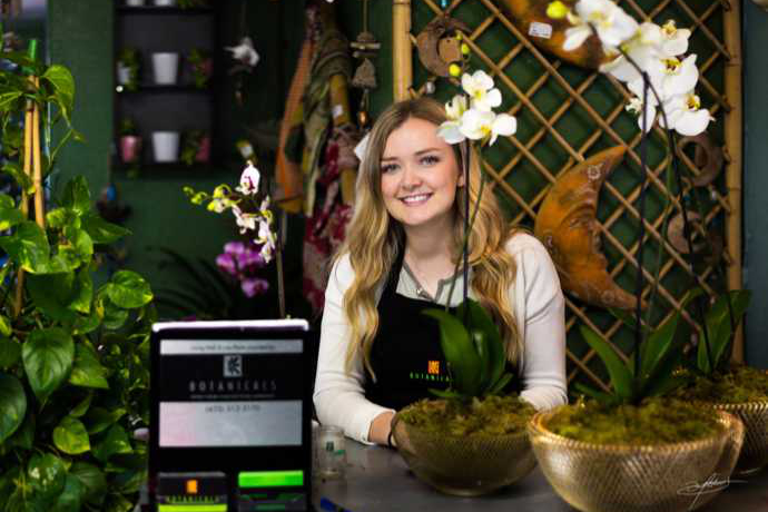 woman smiling and standing behind retail counter with flowers and plants placed on top
